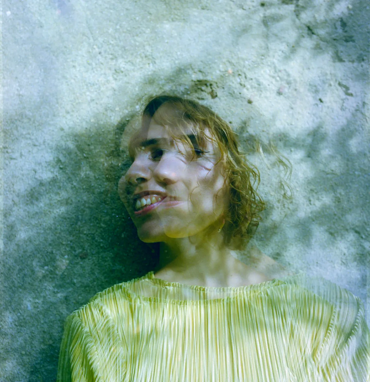 A double-exposed portrait of a smiling person against a mottled green wall.